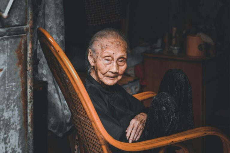 Portrait of an elderly woman sitting in a rocking chair indoors with a contemplative look.
