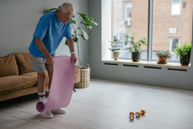 A senior man unfolds a yoga mat indoors, preparing for a fitness routine with dumbbells nearby.