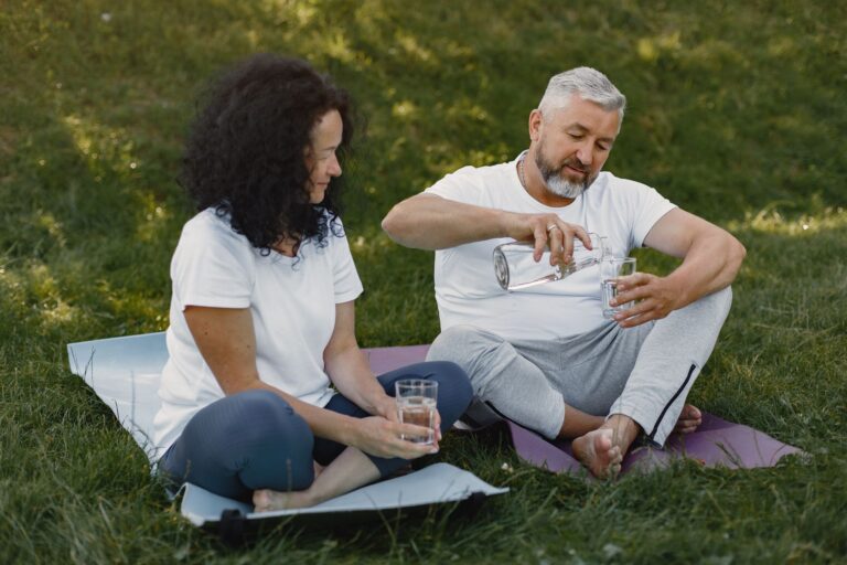 Mature couple sitting on yoga mats outdoors, sharing water and enjoying a peaceful moment.