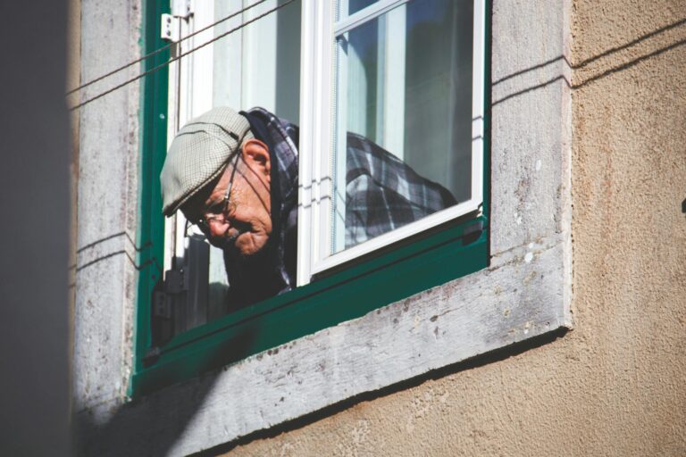 Senior man wearing a beret, peering out a window, showcasing vintage style and contemplation.