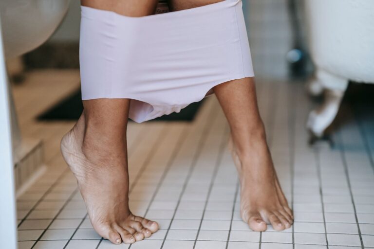 Crop unrecognizable barefoot female in underwear sitting on toilet in bathroom with tiled floor