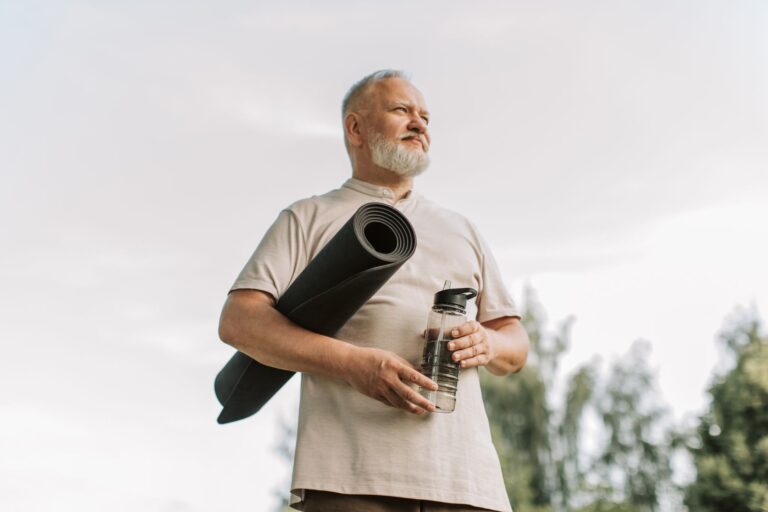 Elderly man with white hair holding a yoga mat and water bottle outdoors.