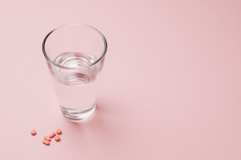 Elevated view of a glass of water next to pink pills on a pastel pink background symbolizing health and wellness.