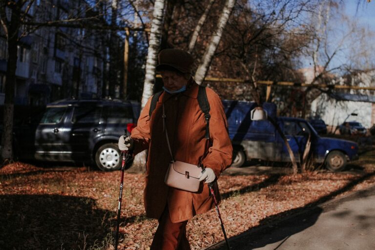 Senior woman using walking sticks in a park with autumn leaves, wearing a brown jacket.