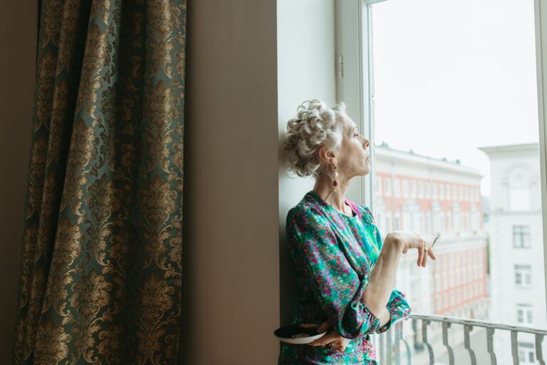 Elderly woman in floral dress smoking by a window indoors.