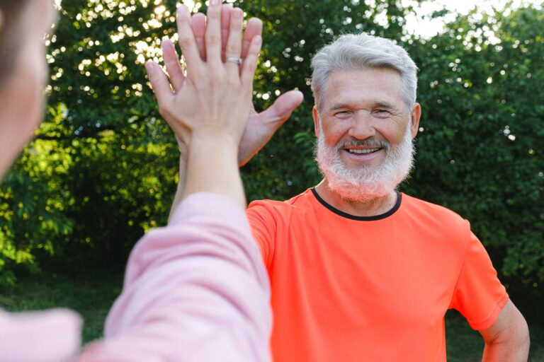 Cheerful aged man in bright clothes smiling and giving high five to friend in sunny day