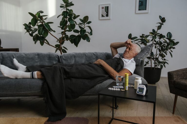 Senior man resting on a couch indoors with medicines on the table, looking unwell.
