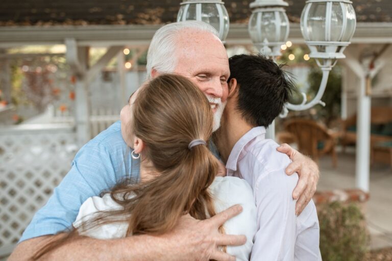 An elderly man joyfully hugs two younger adults in a cozy outdoor setting filled with affection.