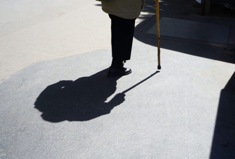 Silhouette of senior walking with a cane on sunlit pavement, highlighting shadows.