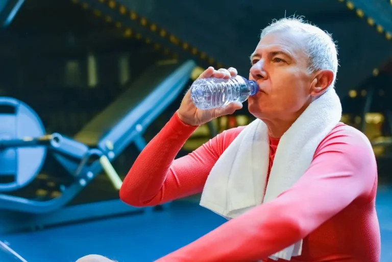 Senior man in gym resting and drinking water post workout for hydration.