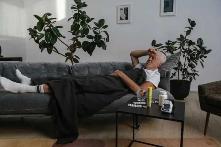 Senior man resting on a couch indoors with medicines on the table, looking unwell.