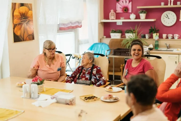 Elderly residents and caregivers enjoying a meal and conversation in a cheerful dining space.