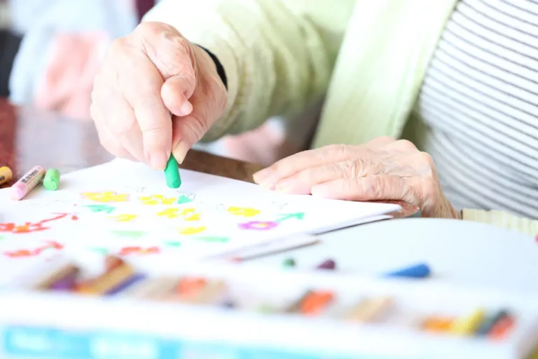 Faceless elderly woman with crooked fingers drawing with crayons at table in nursing home