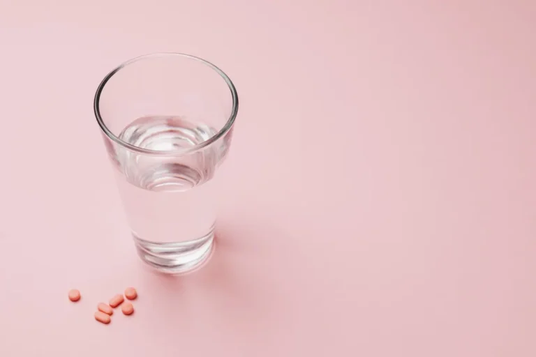 Elevated view of a glass of water next to pink pills on a pastel pink background symbolizing health and wellness.